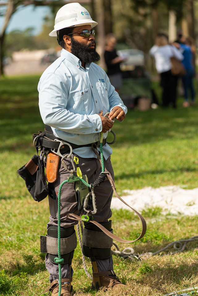 A man with a full beard, sunglasses and a white hard hat puts on safety gear